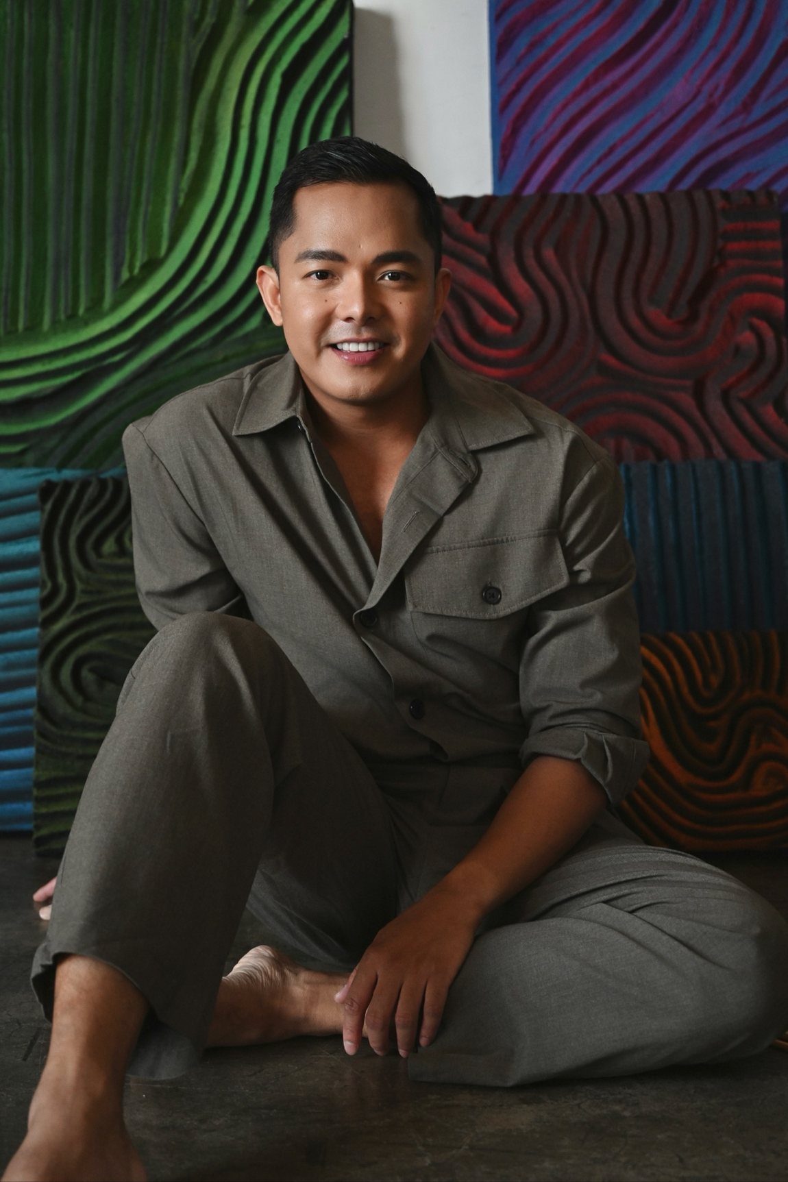 Patrick Viray in a grey jumpsuit, seated on the studio floor smiling in front of his collection of relief panels