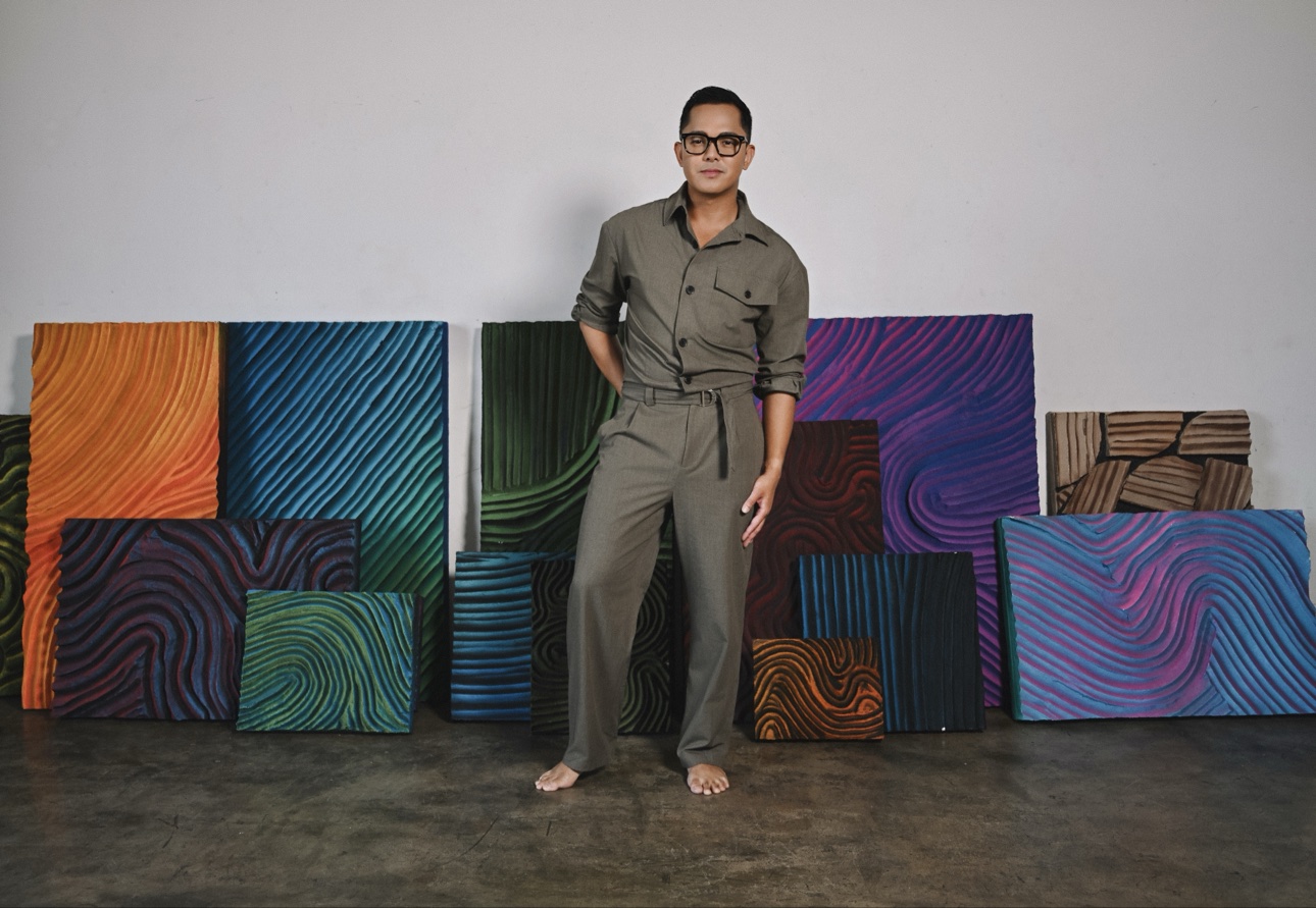 Patrick Viray standing barefoot in his studio in front of a wall of his colorful sculptural relief paintings