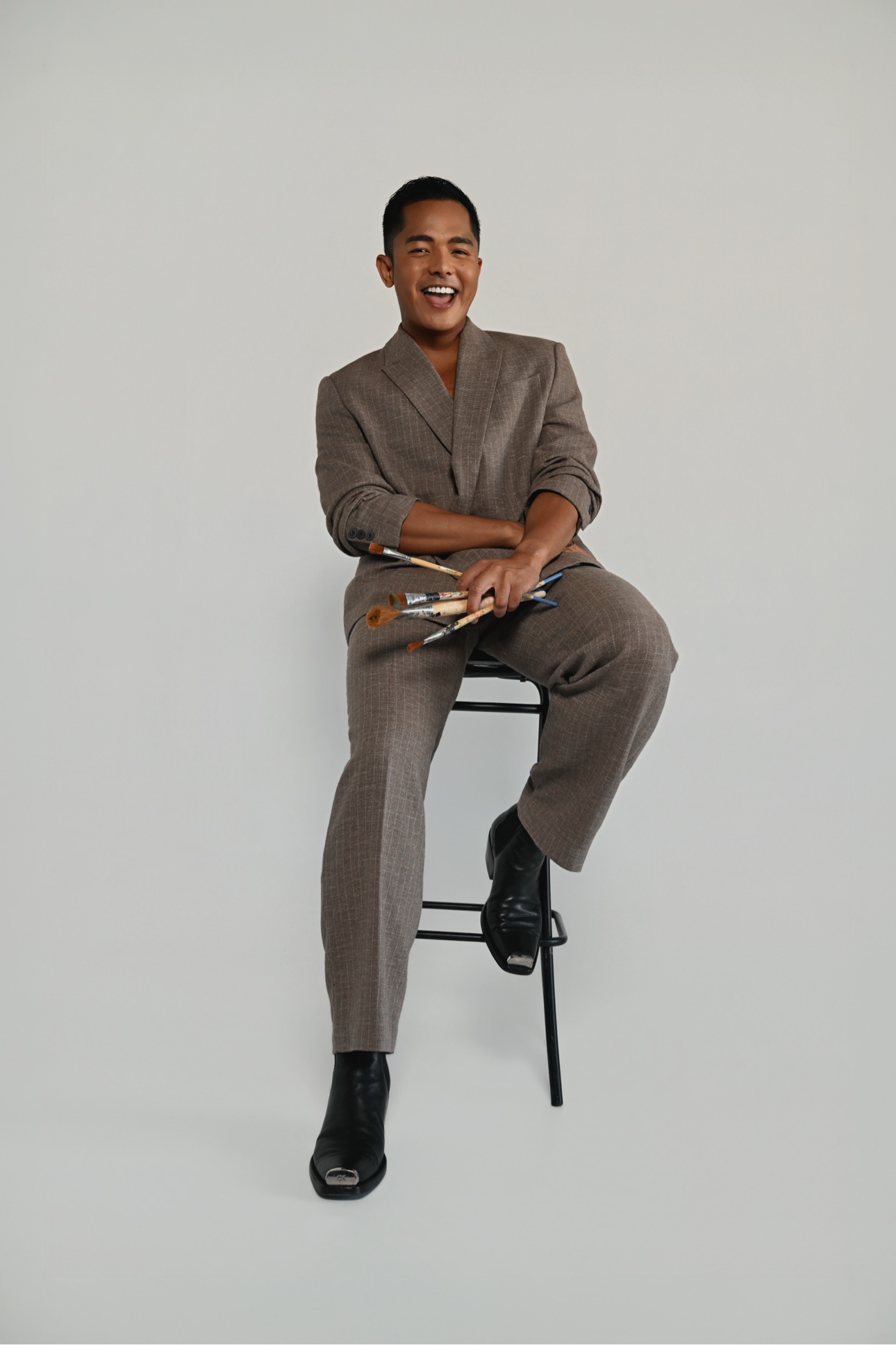 Patrick on a stool laughing, holding a fan of paintbrushes