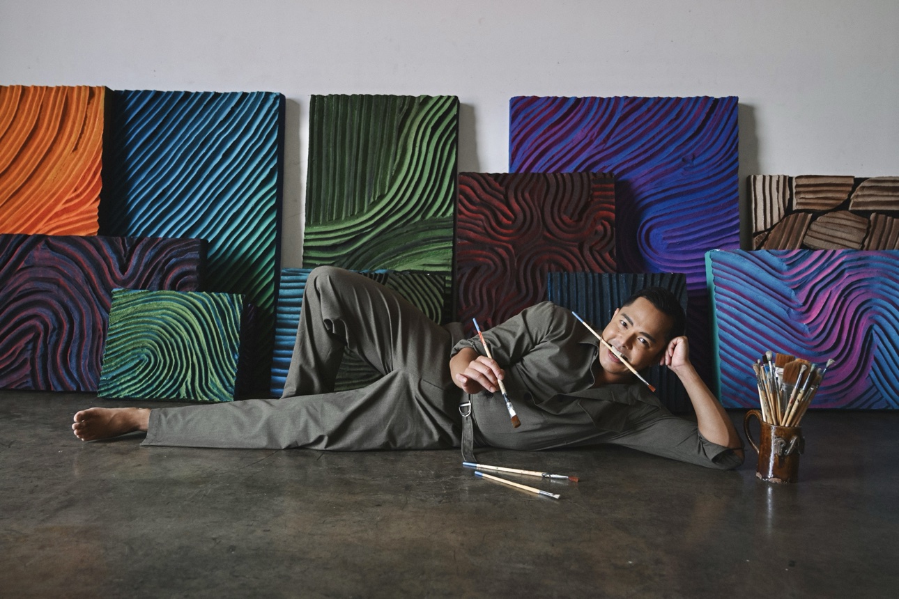 Patrick reclining on the studio floor with paintbrushes, the collection wall behind him