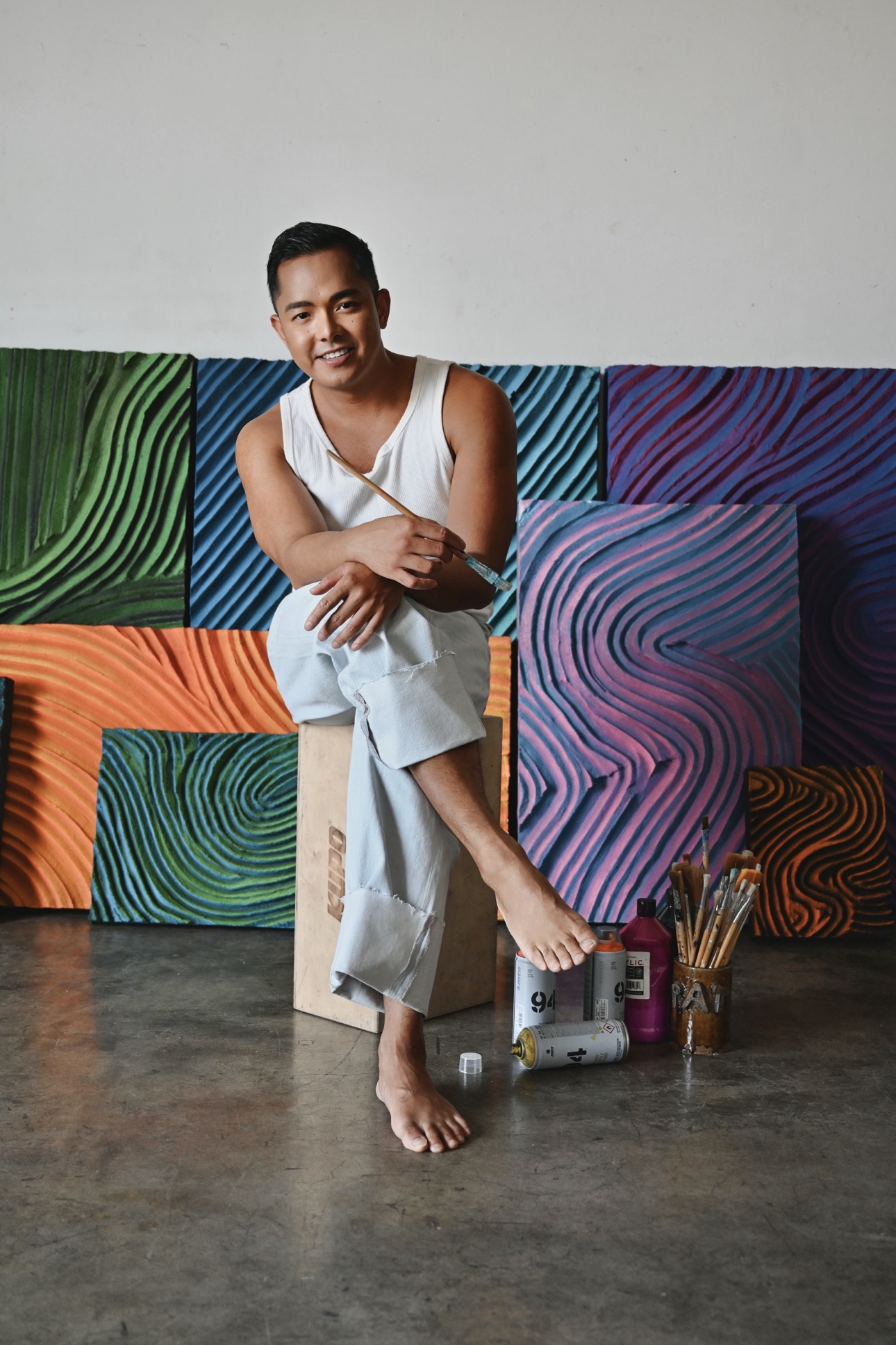 Patrick seated on a wooden cube holding a paintbrush, collection wall behind him