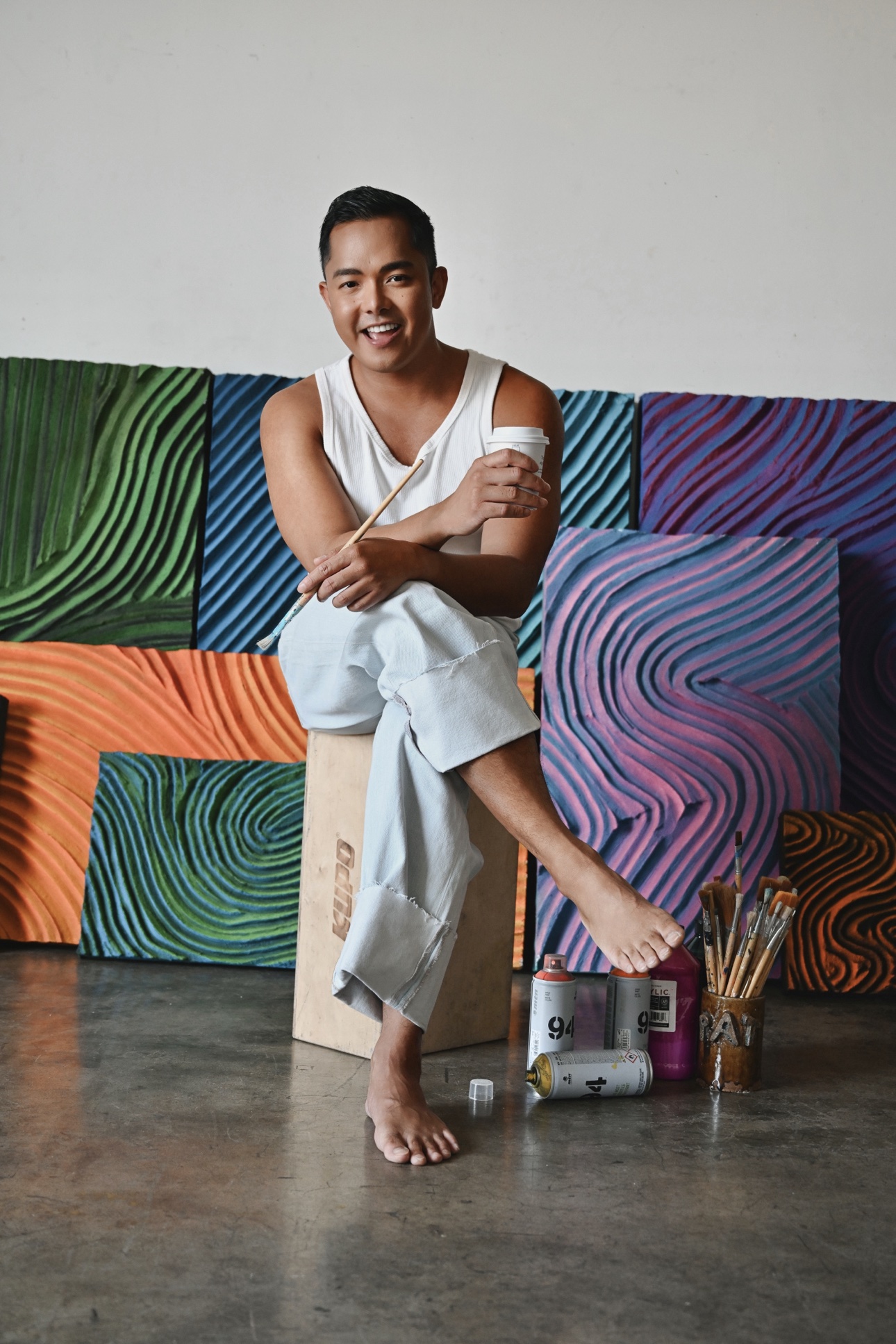 Patrick laughing on a studio block, holding a coffee cup and a paintbrush