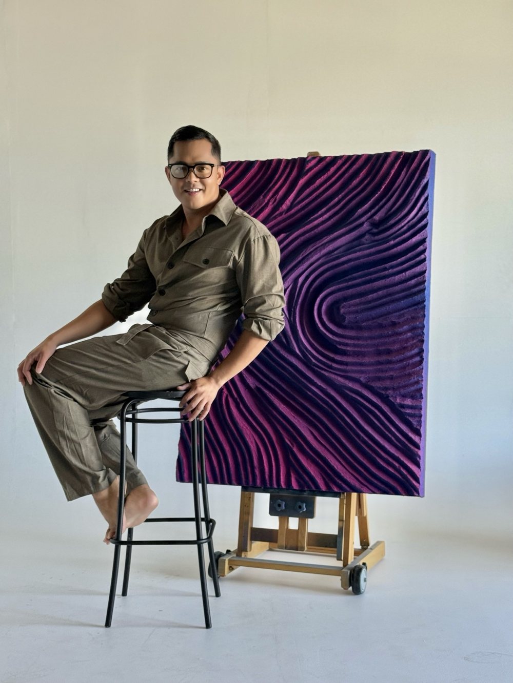 Patrick in a grey jumpsuit and glasses, seated on a stool beside Velvet After Hours on its easel