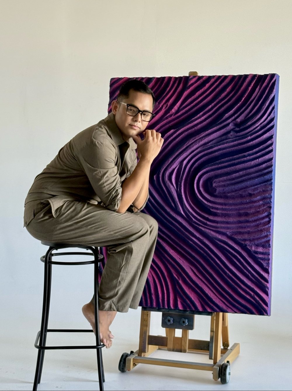 Patrick in a grey jumpsuit and glasses, chin resting on his hand beside the purple relief panel