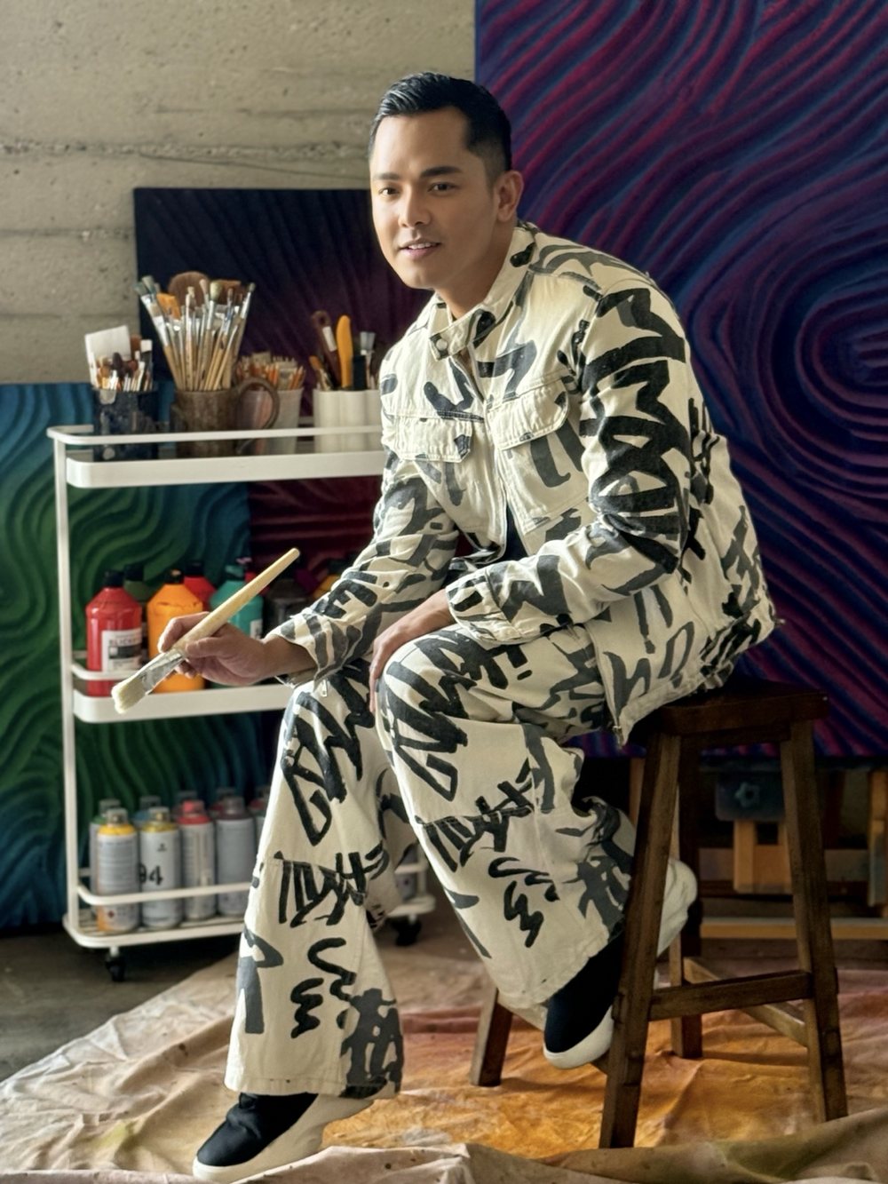 Patrick in the brushstroke print set, seated on a stool in the studio holding a paintbrush